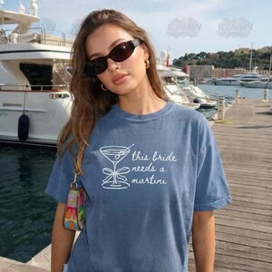 May include: A blue t-shirt with the text "this bride needs a martini" and a martini glass graphic. The shirt is worn by a person wearing sunglasses and gold hoop earrings. The background includes a marina with boats.