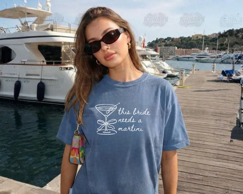 May include: A blue t-shirt with the text "this bride needs a martini" and a martini glass graphic. The shirt is worn by a person wearing sunglasses and gold hoop earrings. The background includes a marina with boats.