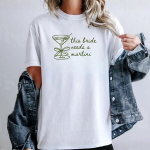 May include: White t-shirt with a green martini glass graphic and the text "this bride needs a martini". The shirt is made of a soft material and is suitable for casual wear.