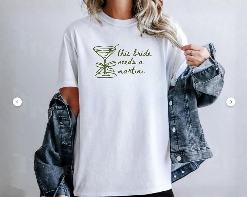 May include: White t-shirt with a green martini glass graphic and the text "this bride needs a martini". The shirt is made of a soft material and is suitable for casual wear.