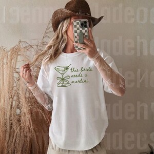 May include: White t-shirt with a green martini glass graphic and the text "this bride needs a martini". The shirt has a round neck and short sleeves. The design is simple and elegant, perfect for a bachelorette party or bridal event.