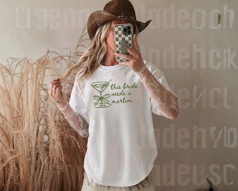 May include: White t-shirt with a green martini glass graphic and the text "this bride needs a martini". The shirt has a round neck and short sleeves. The design is simple and elegant, perfect for a bachelorette party or bridal event.