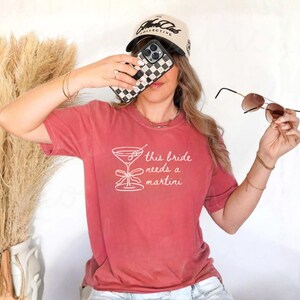 May include: A red-orange t-shirt with the text "this bride needs a martini" in white. The image also shows a black and white checkered phone case, sunglasses, and a baseball cap.