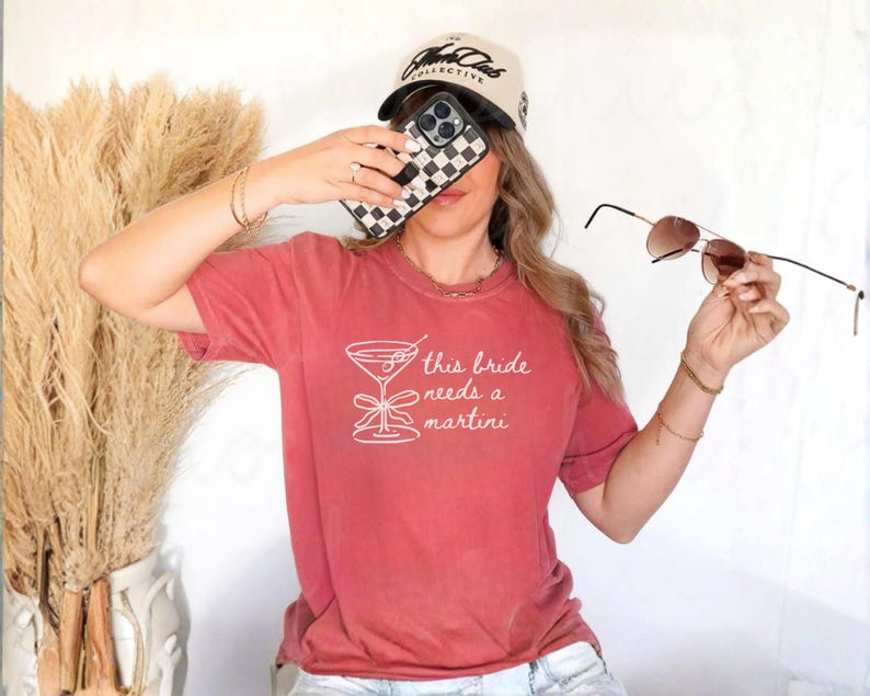 May include: A red-orange t-shirt with the text "this bride needs a martini" in white. The image also shows a black and white checkered phone case, sunglasses, and a baseball cap.