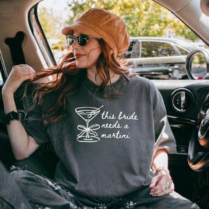 May include: Dark gray t-shirt with the text "this bride needs a martini" and a martini glass graphic. The shirt is worn by a person wearing sunglasses and a brown hat, sitting in a car.