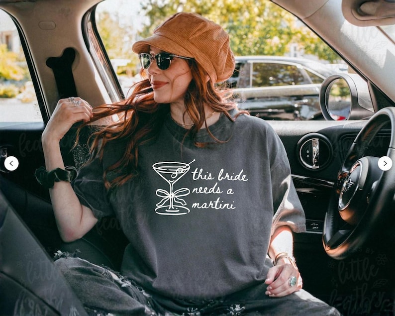 May include: Dark gray t-shirt with the text "this bride needs a martini" and a martini glass graphic. The shirt is worn by a person wearing sunglasses and a brown hat, sitting in a car.