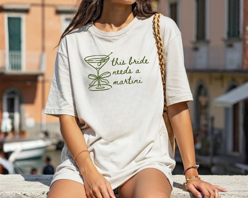 May include: Off-white t-shirt with a green martini glass graphic and the text "this bride needs a martini." The design features a cocktail glass with an olive and a bow. The shirt is a casual, oversized fit.