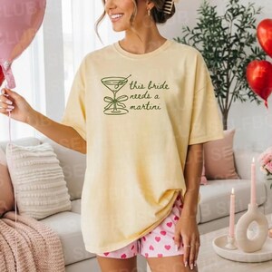 May include: A light yellow t-shirt with the text "this bride needs a martini" in green. The shirt is paired with pink shorts with a heart pattern. Red and pink heart-shaped balloons are in the background.