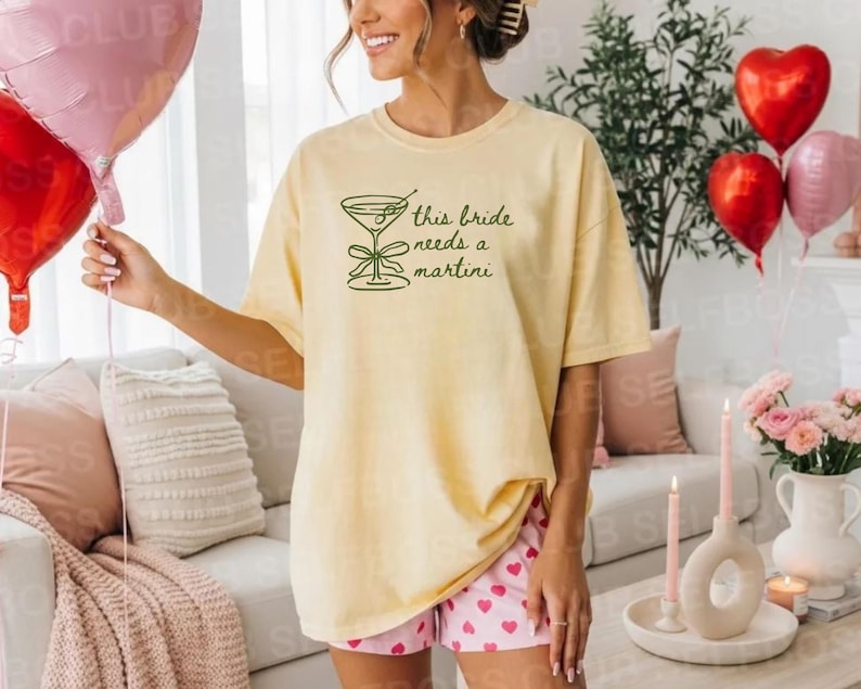 May include: A light yellow t-shirt with the text "this bride needs a martini" in green. The shirt is paired with pink shorts with a heart pattern. Red and pink heart-shaped balloons are in the background.