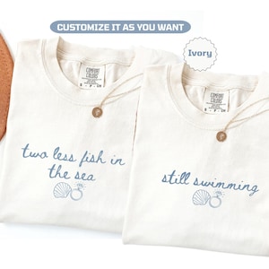 Può includere: Due magliette color avorio con testo blu: "two less fish in the sea" e "still swimming". Ognuna ha una grafica a conchiglia e anello. Collane d'oro con pendenti rotondi sono sulle magliette. Il testo "Customize it as you want" è in alto.