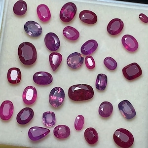 May include: Assortment of polished gemstones in various shapes and sizes, including oval, pear, and rectangular cuts. The gems display shades of red and purple, with variations in clarity and saturation. The gemstones are displayed on a light-colored surface.