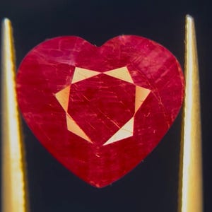 Puede incluir: Una piedra preciosa roja en forma de corazón con una superficie facetada, que muestra un patrón geométrico interno distintivo. La piedra está sujeta por dos pinzas de metal dorado sobre un fondo oscuro, resaltando su color carmesí intenso.