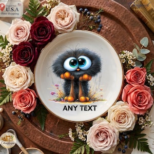 May include: A white ceramic plate featuring a central illustration of a fluffy black creature with large blue eyes and orange feet, surrounded by a decorative arrangement of pink, red, and white roses, green leaves, and the text ANY TEXT.