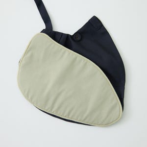 May include: A dark navy blue shoulder bag featuring a distinct asymmetrical, curved silhouette. The front panel is a contrasting light beige, padded fabric with a piped edge. A single dark navy strap and a visible button closure are present.