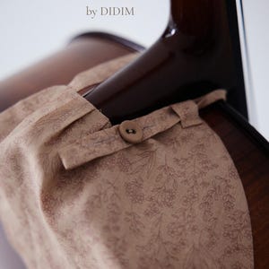 May include: A beige fabric cello drape with a delicate brown floral line-art pattern, featuring a button closure secured around the neck of a dark brown wooden cello. The text CELLO DRAPE by DIDIM is printed in the upper left corner of the image.