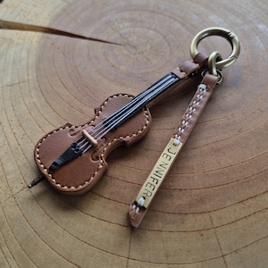 Può includere: Un portachiavi in pelle marrone a forma di violino con corde e ponticello neri. È dotato di un cinturino in pelle con cuciture bianche e una targhetta rettangolare in ottone incisa con il nome JENNIFER, uniti da un anello in ottone.