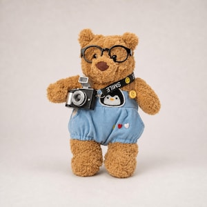 Op de afbeelding: Een bruine teddybeer met een zwarte bril en een blauwe overall. De beer houdt een zwarte camera vast en heeft een band met de tekst "SMILE" en een pinguïn patch. De overall heeft hart- en knoopdetails.