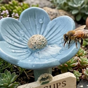 以下が含まれることがあります： 淡い青色の花型のバードバス。中央はテクスチャ加工が施され、縁には蜂がとまっています。バードバスは緑に囲まれ、表面には水滴が付いています。
