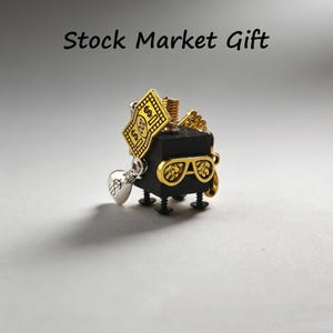 Może przedstawiać: Czarna i złota figurka z okularami przeciwsłonecznymi, znakiem pieniądza i stosu monet. Figurka ma cztery małe nóżki i srebrny urok. Tekst "Stock Market Gift" znajduje się nad figurką.