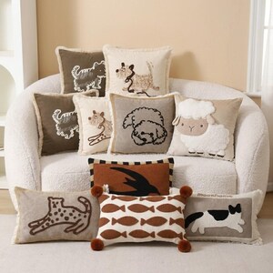 May include: A collection of square and rectangular throw cushions featuring embroidered animal designs including dogs, a sheep, a leopard, and a cat, along with a fish pattern and a bird silhouette, all in neutral earth tones with fringe edges.