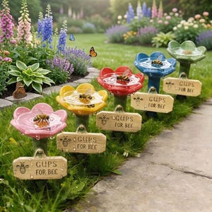 Könnte beinhalten: Eine Reihe von bienenfreundlichen Wasserstellen in Form von bunten Blumen in einem Garten. Jede Wasserstelle hat eine andere Farbe, darunter Rosa, Gelb, Rot, Blau und Grün, und ein kleines Holzschild mit der Aufschrift "GUPS FOR BEE". Bienen sind sichtbar.