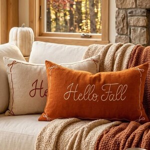 Puede incluir: Un cojín rectangular de color naranja con el texto "Hello, Fall" bordado en blanco en letra cursiva. El cojín tiene un borde fino bordado en blanco con pequeños detalles de lazos en cada esquina, colocado sobre un sofá de color claro.