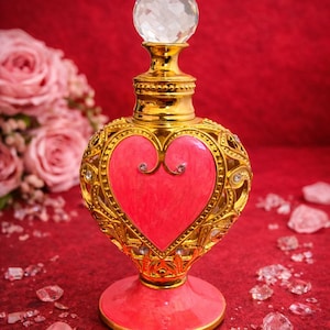 Könnte beinhalten: Dekorative Parfümflasche in Gold mit einem herzförmigen Design in Pink, verziert mit Kristallen. Der Verschluss ist ein facettierter Kristall. Die Flasche steht auf rotem Untergrund. Die Basis ist rosa.