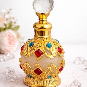 Può includere: Elegante bottiglia di profumo dorata con un tappo trasparente sfaccettato. La bottiglia è adornata con pietre preziose rosse e blu incastonate in un motivo decorativo in filigrana dorata. Anche la base e il collo sono dorati.