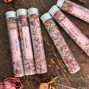Può includere: Diversi tubi di vetro pieni di fiori secchi e sali, etichettati "NEW LOVE". I tubi hanno tappi argentati ed etichette rosa. L'immagine include rose secche e il testo "HANDCRAFTED BATH SOAK • RITUAL SPELL JAR".
