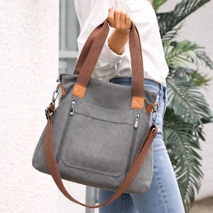May include: Grey canvas tote bag with brown straps and details. Features a front pocket and two zipped compartments. Includes a detachable shoulder strap. The bag is held by a person wearing a white shirt and blue jeans.