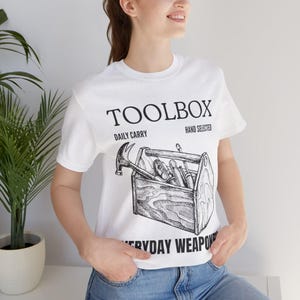 Könnte beinhalten: Weißes T-Shirt mit einer schwarz-weißen Grafik einer Werkzeugkiste voller Werkzeuge. Der Text "TOOLBOX" befindet sich über der Werkzeugkiste, mit "DAILY CARRY" und "HAND SELECTED" auf beiden Seiten. Darunter steht "EVERYDAY WEAPON".