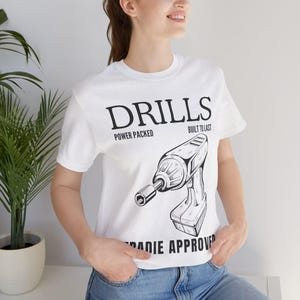 以下が含まれることがあります： 白いTシャツに、ドリルの黒いグラフィックと、その上に「DRILLS」の文字が描かれています。「POWER PACKED」と「BUILT TO LAST」の文字がドリルの画像の上にあります。「RADIE APPROVED」の文字が下にあります。