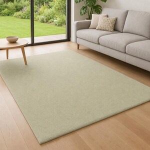 May include: A rectangular area rug in a solid light sage green color with a short, dense pile texture. The rug is positioned on a light-toned wooden floor, extending from the foreground toward a gray sofa and a small round wooden side table.