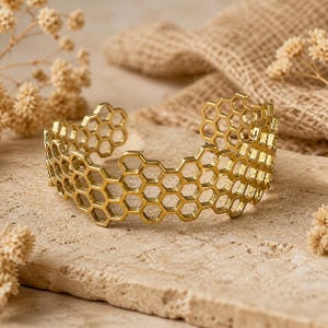 Könnte beinhalten: Goldfarbenes Armreif-Armband mit Wabenmuster. Das offene Armband besteht aus Metall und weist ein sich wiederholendes Muster aus sechseckigen Formen auf. Das Armband wird auf einer hellen Steinoberfläche präsentiert.