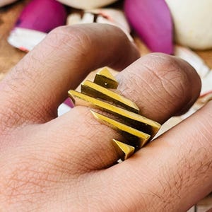 Könnte beinhalten: Ein goldfarbener Ring mit geometrischem Design. Der Ring hat mehrere dreieckige und rechteckige Formen. Der Ring wird an einem Finger getragen und zeigt seine kühne und moderne Ästhetik. Das Design des Rings ist eckig und auffällig.