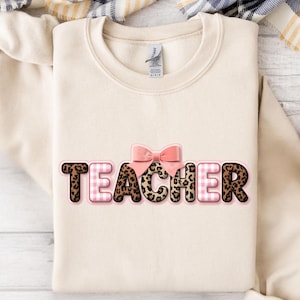Puede incluir: Una sudadera color crema con la palabra "TEACHER" en un diseño divertido. Las letras combinan estampado de leopardo, cuadros rosas y contornos rosas. Un lazo rosa se encuentra encima de la palabra, agregando un toque decorativo. La sudadera está doblada sobre una superficie neutra.