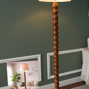 May include: A tall, wooden floor lamp with a light beige lampshade. The lamp's stem is made of stacked, rounded wooden beads. The lamp is set against a dark green wall, with a glimpse of a living room in the background.