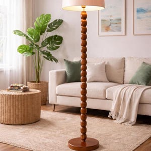 May include: A wooden floor lamp with a beige lampshade. The lamp has a dark brown, beaded column and a round base. The lamp is in a living room with a sofa, rug, and plants.