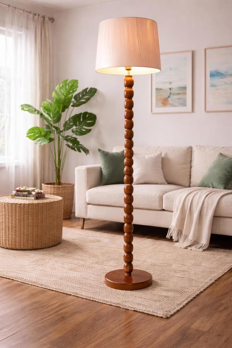May include: A wooden floor lamp with a beige lampshade. The lamp has a dark brown, beaded column and a round base. The lamp is in a living room with a sofa, rug, and plants.