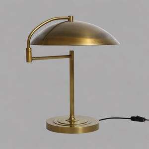 May include: A brass desk lamp with a curved arm and a domed shade. The lamp has a round base and a straight vertical support. The lamp is plugged into a wall outlet with a black cord.