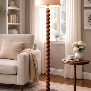 May include: A tall, wooden floor lamp with a beige lampshade. The lamp has a series of round, wooden beads along its stem. The lamp is placed on a round, wooden base. The lamp is in a living room setting.