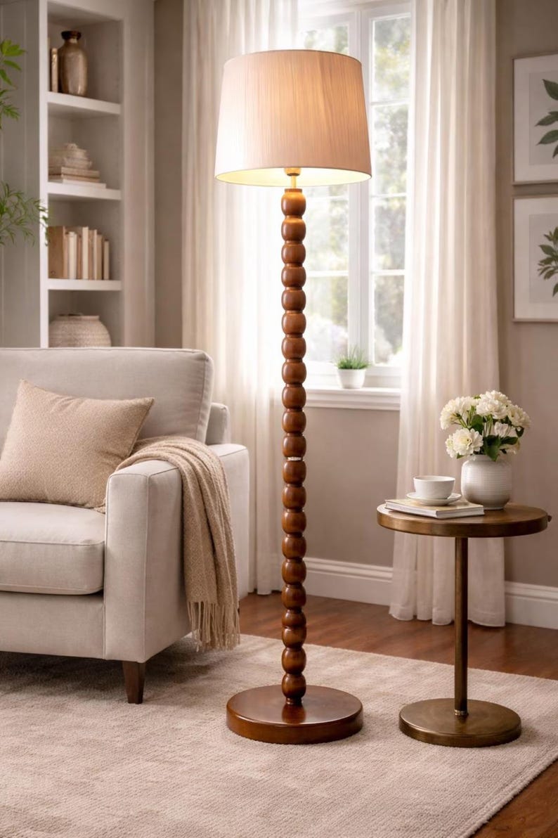 May include: A tall, wooden floor lamp with a beige lampshade. The lamp has a series of round, wooden beads along its stem. The lamp is placed on a round, wooden base. The lamp is in a living room setting.
