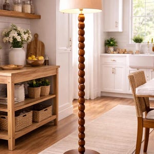 May include: A wooden floor lamp with a beige lampshade. The lamp has a dark brown, beaded stem and a round base. The lamp is in a kitchen with wooden shelves, a table, and a rug.