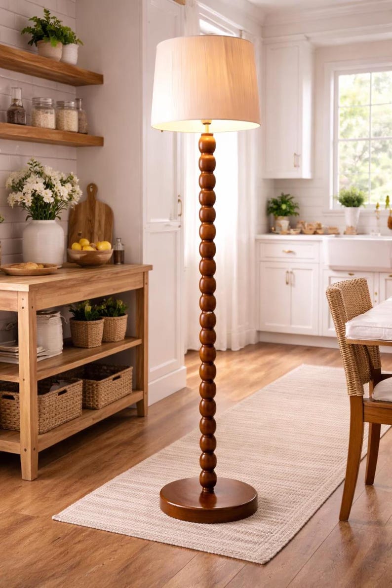 May include: A wooden floor lamp with a beige lampshade. The lamp has a dark brown, beaded stem and a round base. The lamp is in a kitchen with wooden shelves, a table, and a rug.