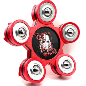 May include: A five-pointed star-shaped red fidget spinner with black accents and five silver-colored metal ball bearings. The center features a circular black inlay with a graphic illustration of a masked character and the text The Villains.