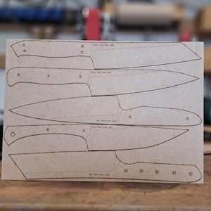 Könnte beinhalten: Ein rechteckiges Stück Pappe mit Messerschablonen. Die Vorlagen sind schwarz umrandet und zeigen verschiedene Messerarten. Der Text "diy-knives.de" ist auf der Pappe aufgedruckt. Der Hintergrund ist eine verschwommene Werkstatteinstellung.