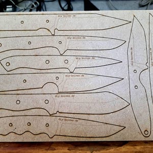 Peut inclure: Une plaque de bois brun clair avec des contours de couteaux découpés au laser. Le texte "diy-knives.de" est visible. Les couteaux ont des formes variées et des trous pour les manches.