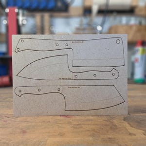 Könnte beinhalten: Eine rechteckige hellbraune Faserplatte als Schablone mit drei lasergebrannten Messerkonturen. Die Designs umfassen ein Hackmesser, ein Kochmesser und ein kleineres Hackmesser, jeweils mit dem Text diy-knives.de am Griffbereich.