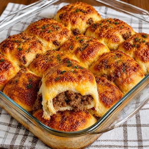 May include: A glass baking dish filled with baked, golden-brown rolls. The rolls are topped with melted cheese and a sprinkle of green herbs. One roll is broken open, revealing a savoury meat filling. The dish sits on a checkered cloth.