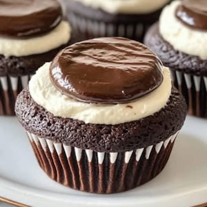 Può includere: Un cupcake al cioccolato con una base di torta marrone scuro in un pirottino di carta plissettato, sormontato da uno spesso strato di glassa bianca e un disco liscio e circolare di ganache al cioccolato fondente centrato, con altri cupcake.